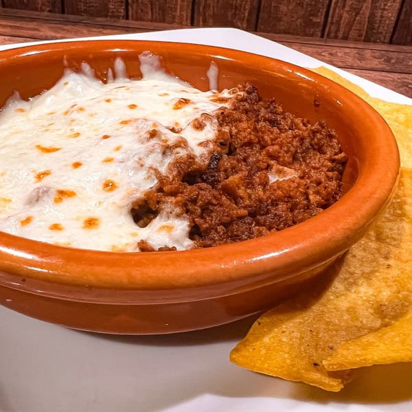 Melted white cheese over chorizo in an orange bowl, with tortilla chips on the side.