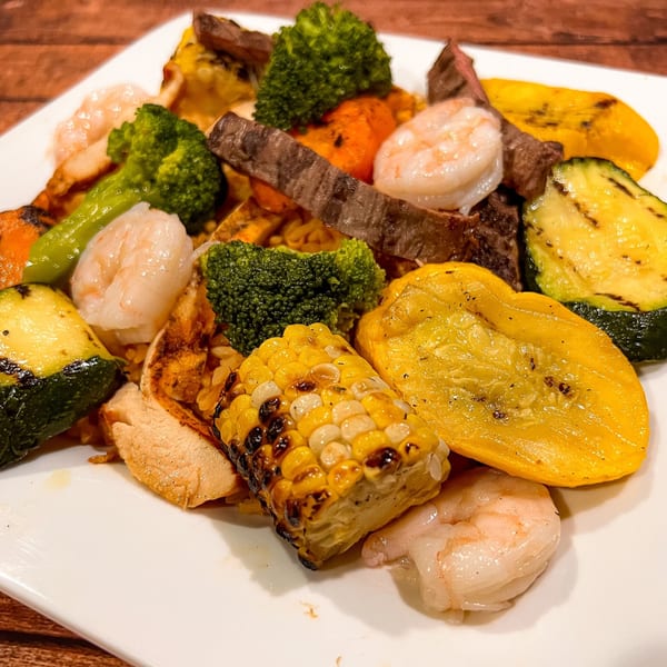Grilled shrimp, steak, chicken, corn, zucchini, yellow squash, and broccoli on a white plate.