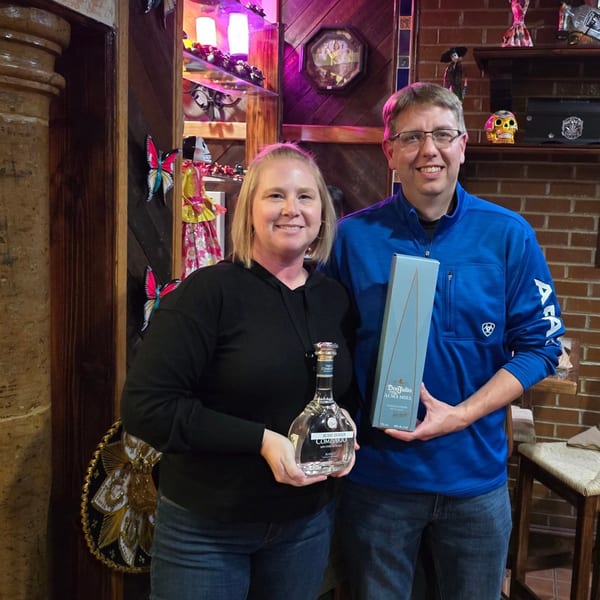 Two smiling people holding bottles of tequila, a woman with Comisario and a man with Don Julio.