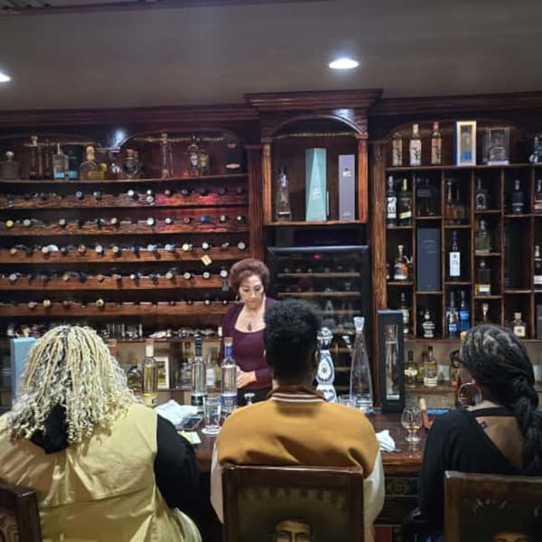 A woman behind a bar with shelves of tequila bottles, serving three customers at Casa Juarez.