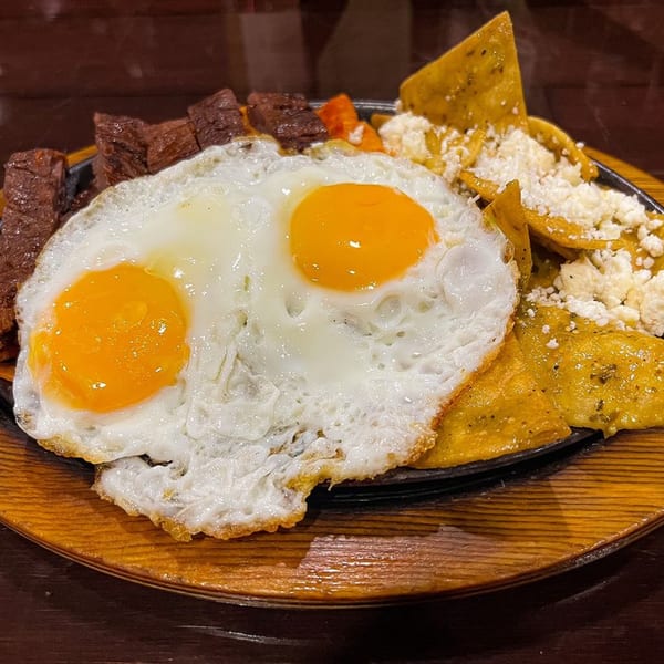 Two sunny-side-up eggs, steak, and chilaquiles with cheese on a sizzling platter.