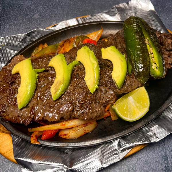 Sizzling Mexican steak fajitas with avocado slices, jalapeños, and lime on a hot skillet.