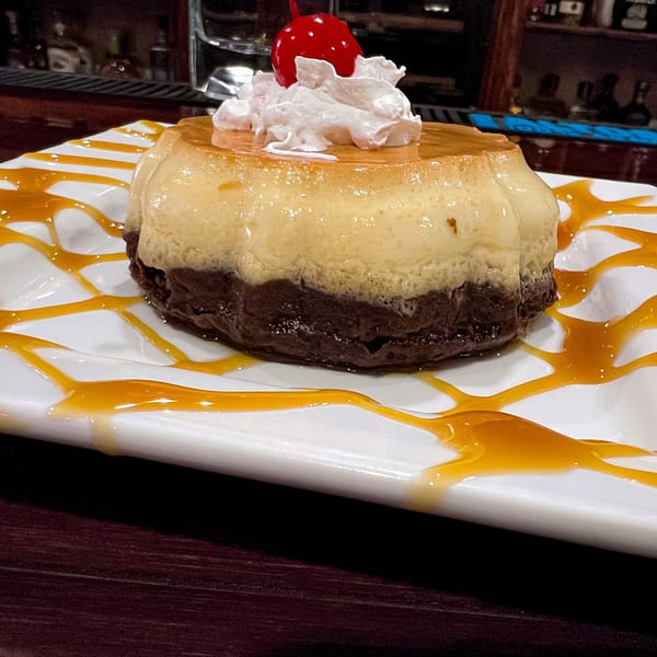 Flan dessert with chocolate base, whipped cream, and a cherry, drizzled with caramel on a white plate.